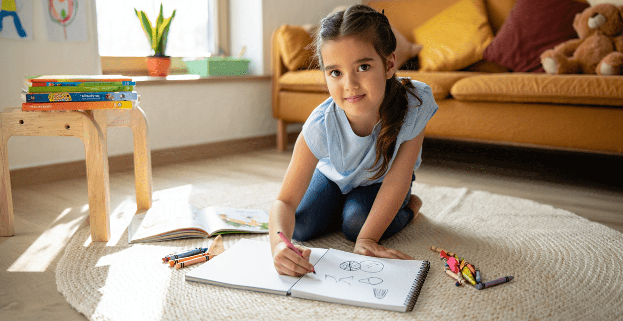 Child drawing in a sketchbook with crayons on a rug.