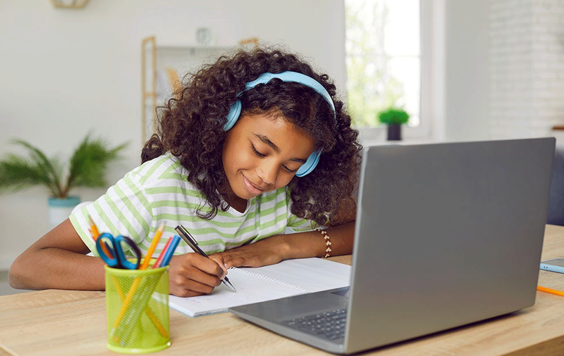 Child with headphones writing in notebook next to laptop.