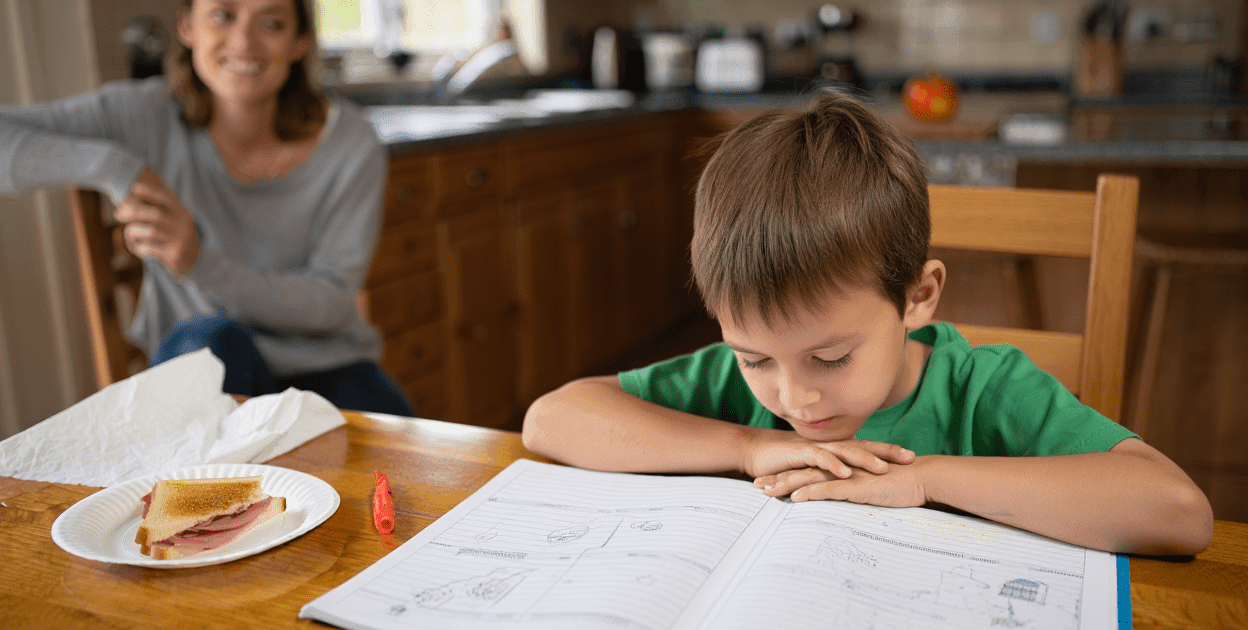 Child studying at a table with an adult in the background.